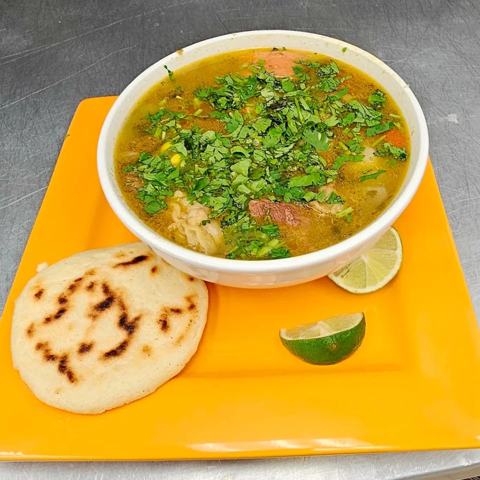 Hearty Mexican soup with meat, vegetables, and cilantro, served with a tortilla and lime wedges.