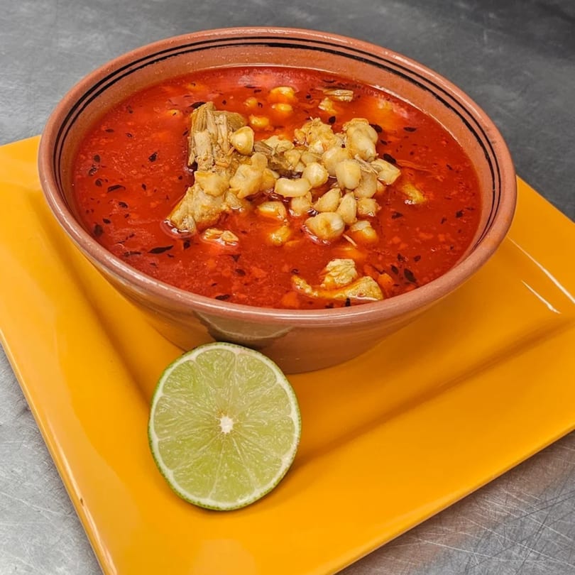A bowl of Mi Tierra's red pozole with hominy and pork, garnished with a lime slice.